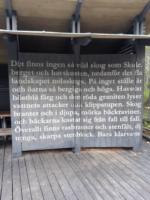 Träkonstruktion med text tryckt på fasaden som beskriver en naturupplevelse, omgiven av trägolv och tak.