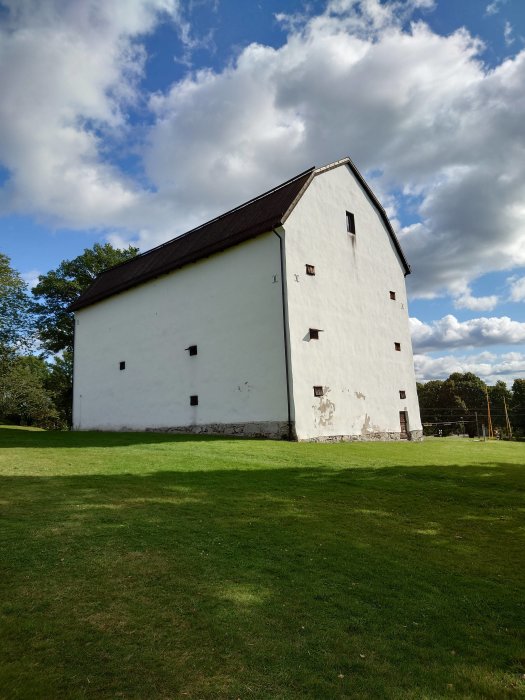 Vitputsad historisk byggnad med brunt tak under en molnig himmel omgiven av en grön gräsmatta.