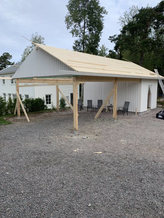 Underkonstruktion av carport med limträpelare, plintar och påbörjat råspont-tak mot hus och träd.