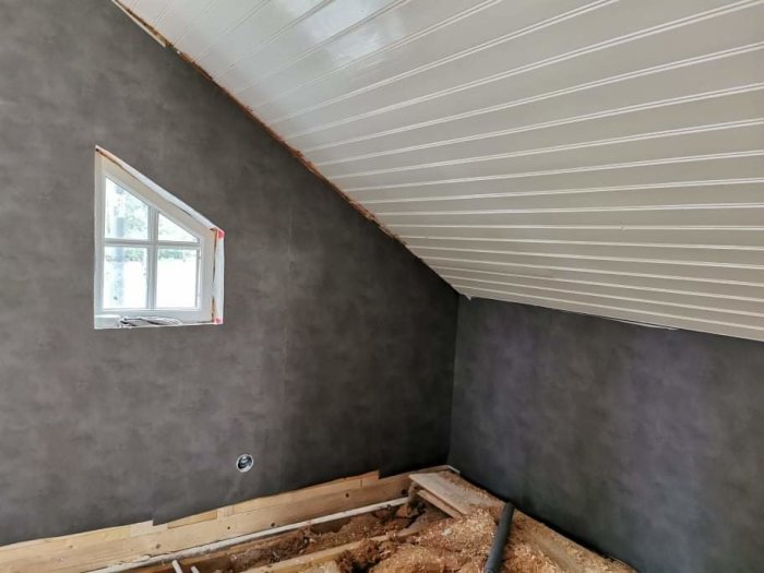 Ett rum med grå kalktapet på snedtak och vägg med fönster, vitt innertak och synligt golvbjälklag.