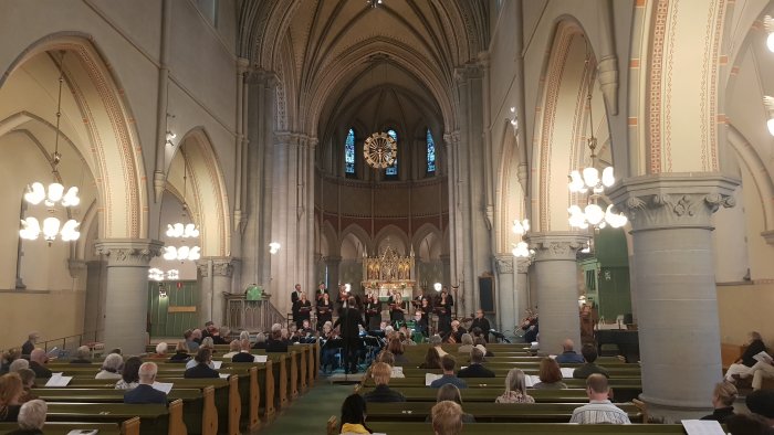 Körframträdande i kyrka med publik i bänkar och färgglatt fönster i fonden.