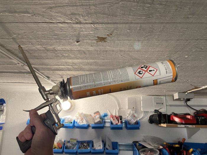 Hand håller en skumgummi-pistol med fogskumstuber riktad uppåt mot ett träbjälklag med verktyg i bakgrunden.