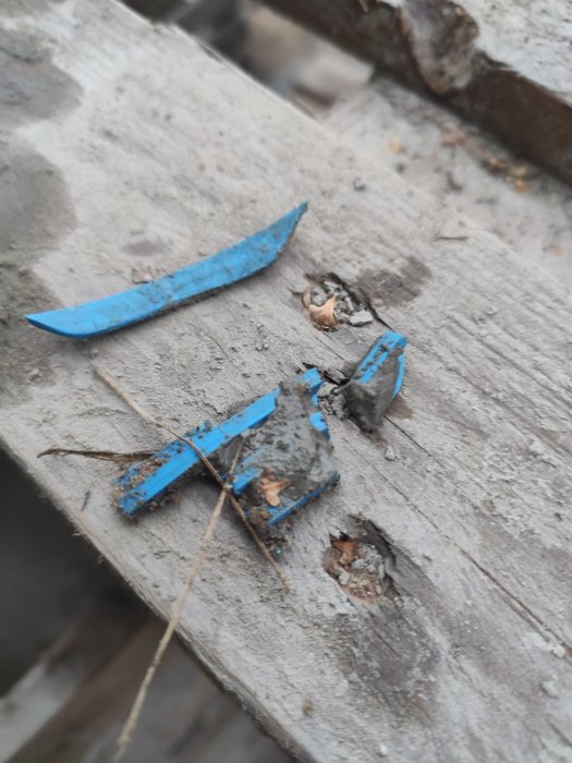 Blå trasiga plastbitar och småstenar på en brun träyta, möjlig skada från borrningsutrustning.