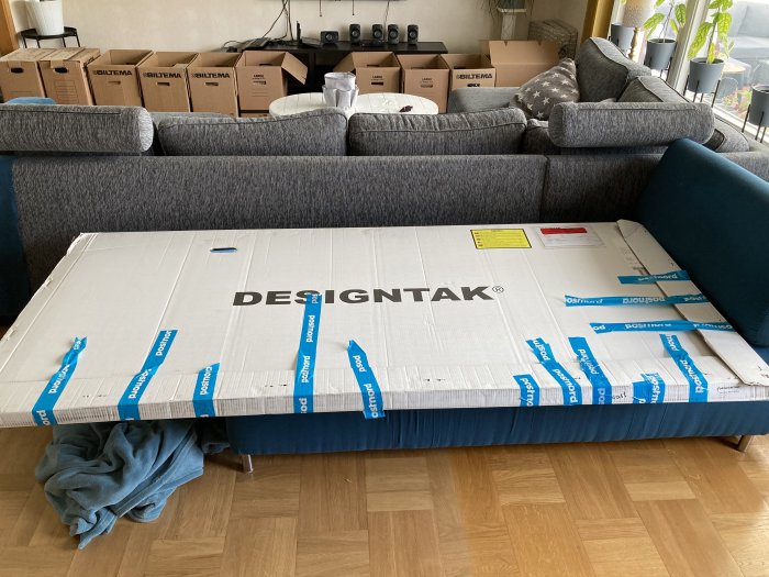 Ett stort paket från Designtak placerat i ett vardagsrum med flyttkartonger i bakgrunden.