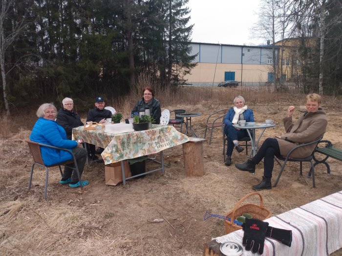 En grupp människor som sitter vid ett improviserat bord utomhus på en tom tomt med vegetation i bakgrunden.