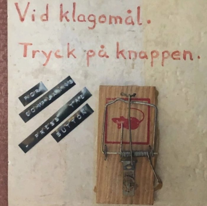 Skylt med texten "Vid klagomål. Tryck på knappen." ovanför en råttfälla, ironisk referens till klagomålshantering.