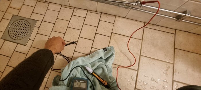 Hand håller i en spänningsprovare mot ett vattenrör i ett badrum, med multimeter som visar 170v och en handduk på golvet.