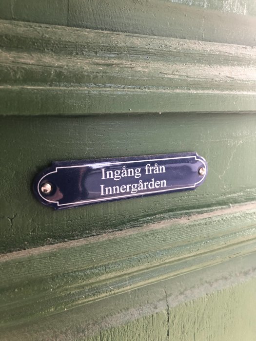 Blå emaljskylt med texten "Ingång från Innergården" monterad på en grön trävägg.