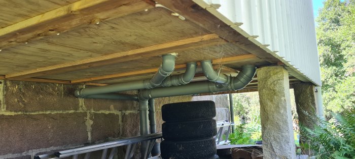 Oisolerade vattenledningsrör under trägolvet i ett utrymme med synliga stenväggar och stödpelare.