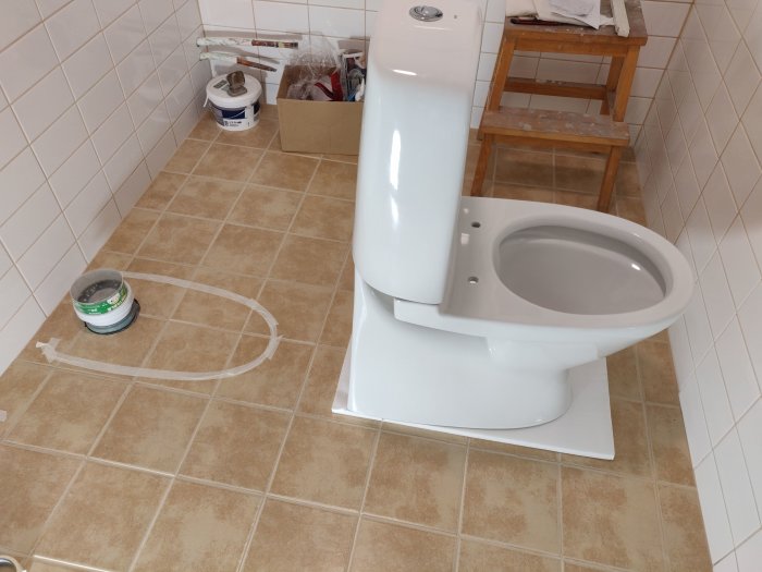 Toalett provplacerad på badrumsgolv med markerade mått för installation, rör och verktyg synliga i bakgrunden.