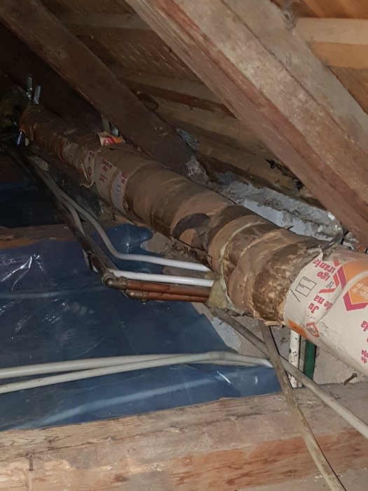 Gamla isolerade rör tätt intill varandra på ett vindsgolv med träbjälkar och sliten isolering.