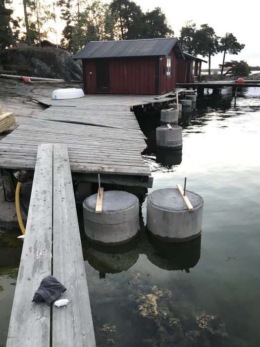 Träbrygga med betongflottar framför röd sjöbod vid skymning, reparation pågår, verktyg synligt.