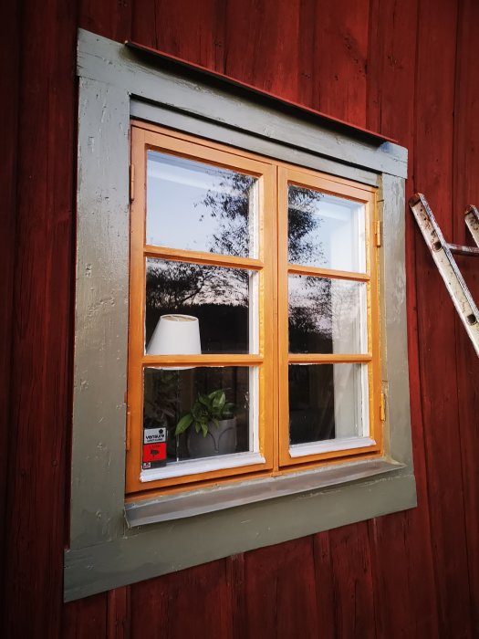 Nyinstallerat fönster med träram på en rödmålad stuga, målararbete pågår, stege skymtar till höger.