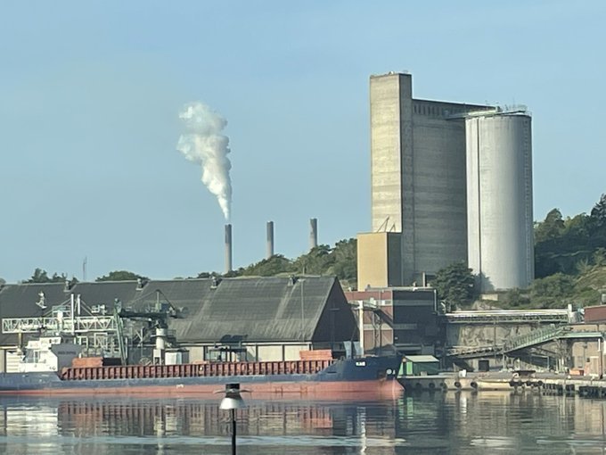 Karlshamnsverket med rök som stiger upp från skorstenen vid kajen, med blå himmel i bakgrunden.