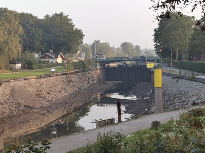 Torrlagd sluss med öppen vägbro och reflekterande vatten, omgiven av träd och en tätbebyggd väg.