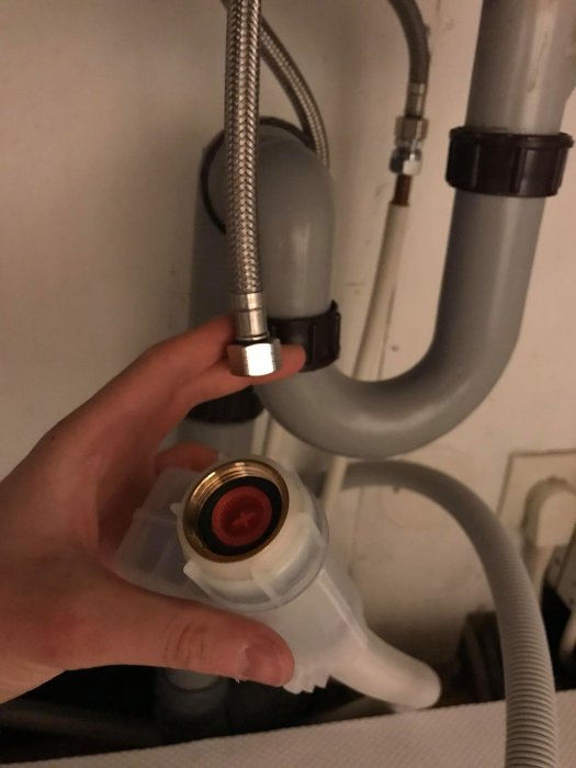 Hand håller en vit diskmaskinsanslutning med en metallände, bredvid gråa avloppsrör under diskbänk.
