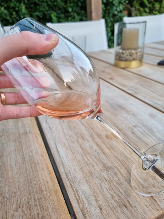 Hand som håller ett glas rosévin över ett träbord utomhus.