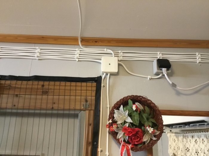 Elledningar längs vägg, en router, och en väggkorg med konstgjorda blommor inomhus.