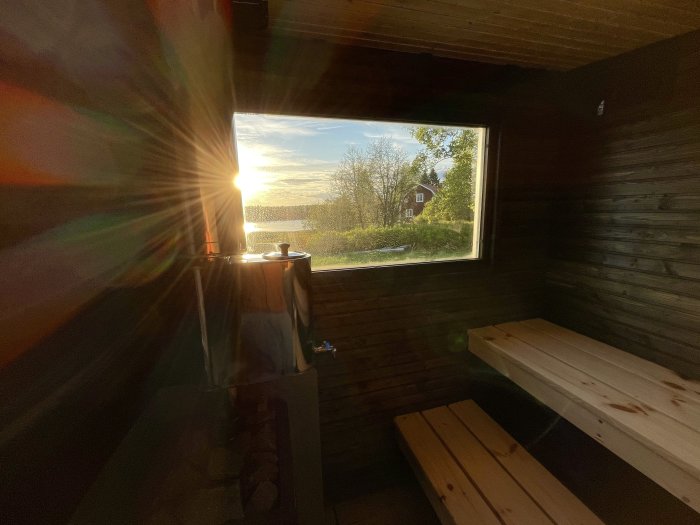 Solnedgång genom fönster i bastu med träbänkar och grönt landskap utanför.