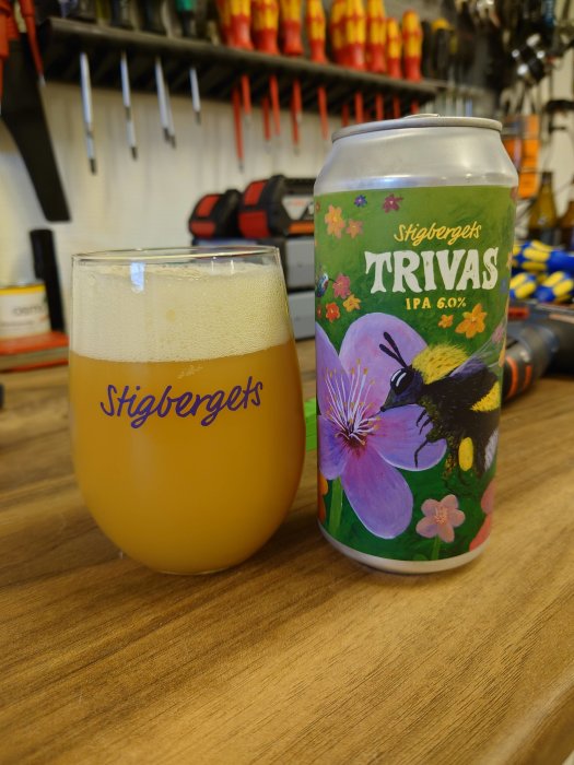 Ölglas märkt med "Stigbergets" och ölburk "Stigbergets TRIVAS IPA" på ett träbord med verktyg i bakgrunden.