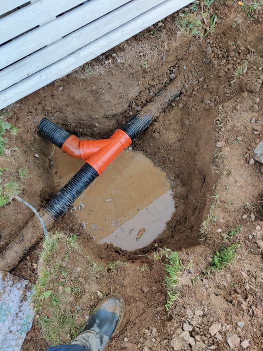 Grävd grop med orange konnektor sammanfogande dräneringsrör fylld med vatten nära husgrund.