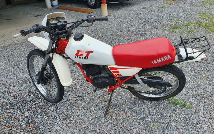 Röd och vit Yamaha DT50 moped parkerad på en grusunderlag.