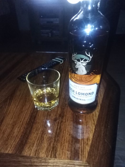 Flaska av Loch Lomond whisky och ett whiskeyglas halvfullt på ett mörkt träbord.