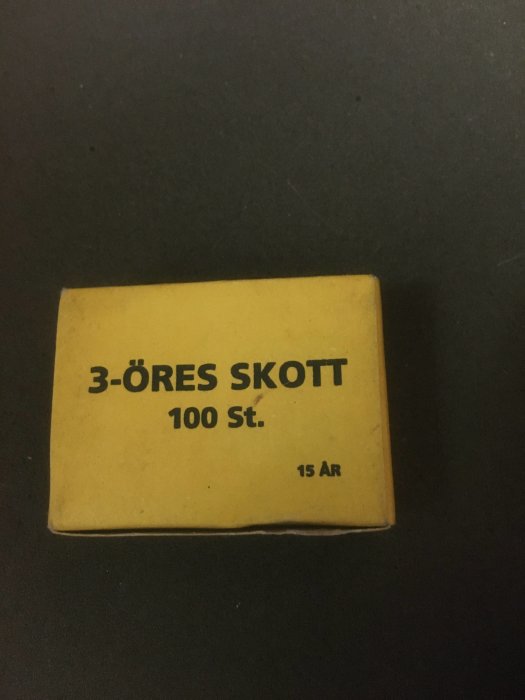 Gammal förpackning märkt "3-ÖRES SKOTT 100 St." som ligger på en mörk yta.