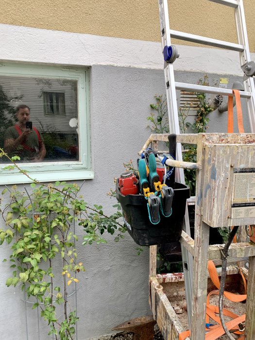 Verktygshink hängande på stege vid en husvägg med reflektion av person som fotograferar i fönstret.