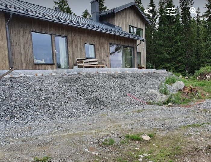 Plats för anläggning av stödmur med markerat område framför trähus och grusat underlag.