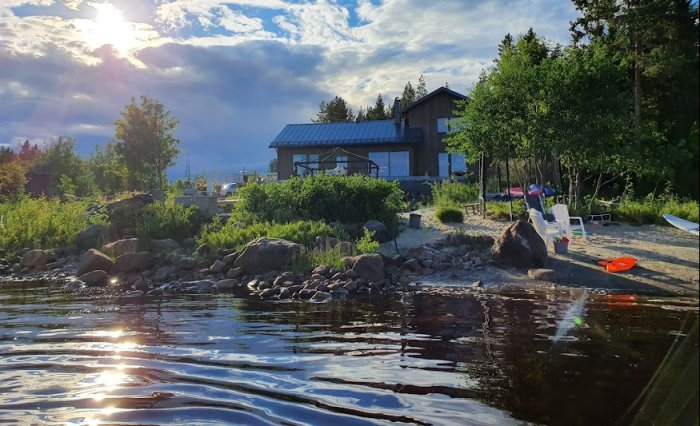 Vy över strandtomt med hus, trädgård och sjö under solnedgång, planeras för stödmur.
