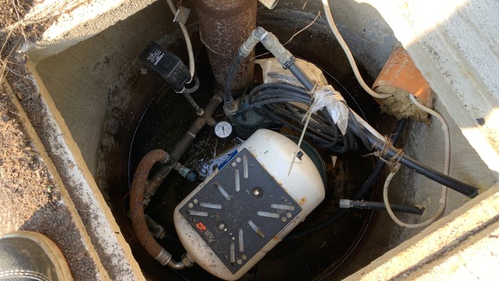 Bild på en brunn med vattenpump, tryckströmbrytare, rör och elektriska kablar, allt ser gammalt och slitet ut.