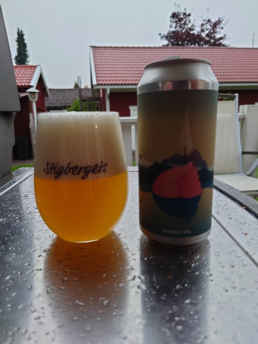 Ölglas fyllt med öl och en burk dubbel IPA på ett vått bord utomhus, med röda hustak i bakgrunden.