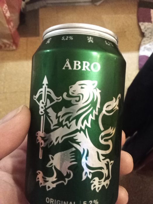 En hand håller en grön ölburk med logotypen och namnet på ett svenskt ölbryggeri, 5,2% alkohol.