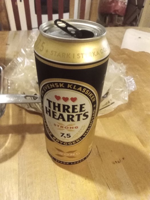 Öppnad ölburk av märket Three Hearts Strong med 7,5 % alkoholhalt placerad på ett träbord.