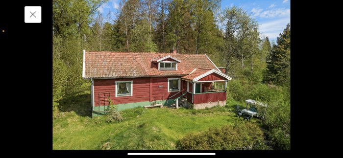 Rött hus med grönt tak i soligt landskap, omgivet av träd och grön gräsmatta med utemöbler framför.