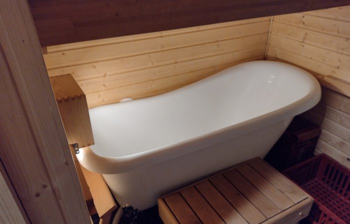 Vintagestil badkar med lång duschslang i ett trärums hörn, röda lådor under för vedförvaring.