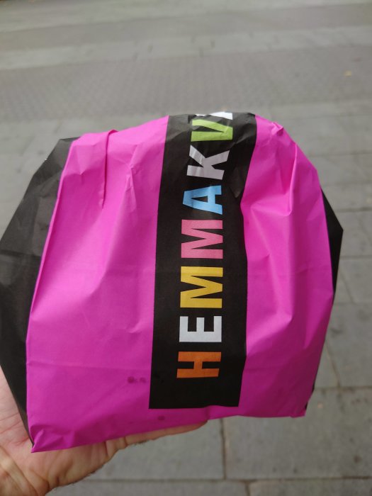 Hand håller en rosa och svart papperskasse med texten "HEMMAK" tryckt på sidan.