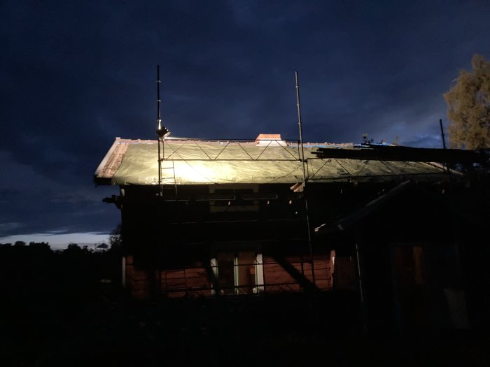 Täckt takkonstruktion på hus med presenning vid skymning, ställningar runt huset.