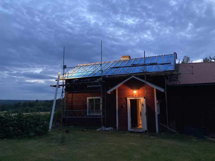 Hus under renovering med halvt färdigt tak och blå presenning, ställning vid skymning.