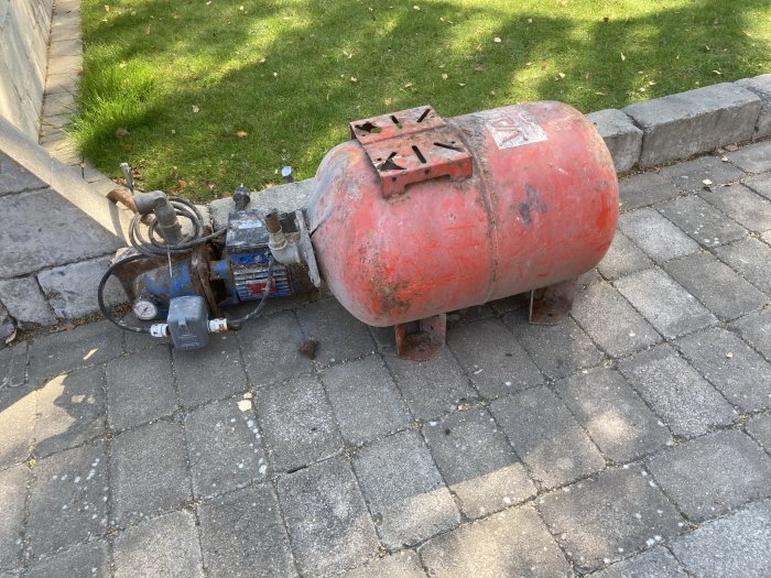 En rostig, gammal hydroforpump på stenplattor med gräs i bakgrunden.