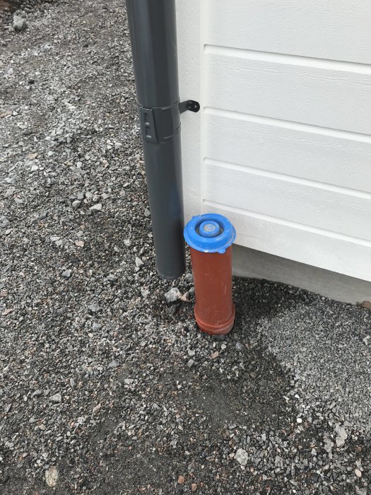 Stuprör på en vit vägg bredvid ett dagvattenrör med blått lock i marken.