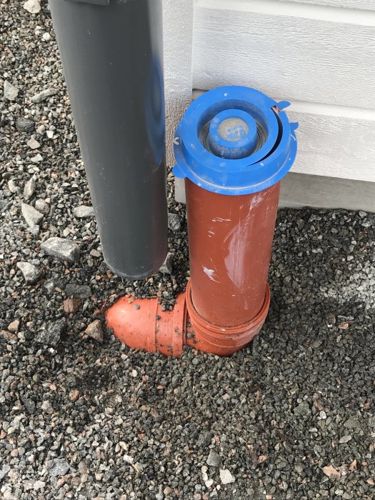 Avloppsrör i marken med en anslutning till ett stående grått rör och en blå kork på toppen.