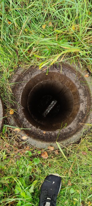 Öppen dagvattenbrunn i gräsmark med synlig fot i nedre högra hörnet som indikerar inspektion eller tester.