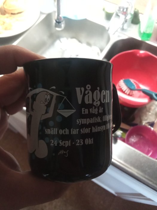 Svart mugg med stjärnteckenmotiv för Vågen och text om egenskaper hålls framför diskho.