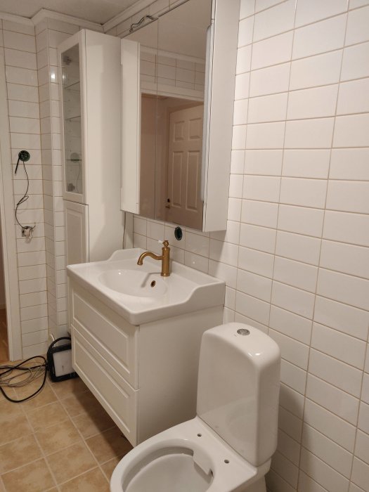 Renoverat badrum med toalettstol, vit handfatsskåp med mässingskran och spegelskåp på kakelvägg.