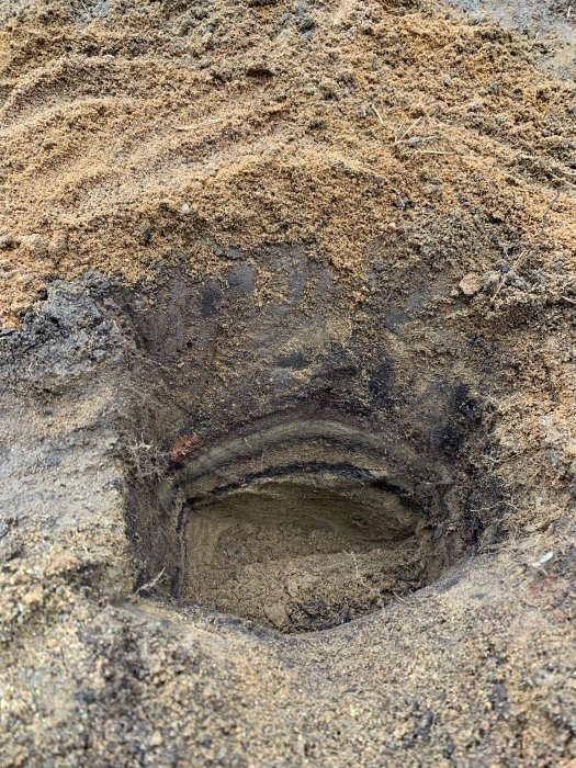 Närbild på ett grävt hål som visar olika jordlager med sand och mörkare jord i en grävd yta.