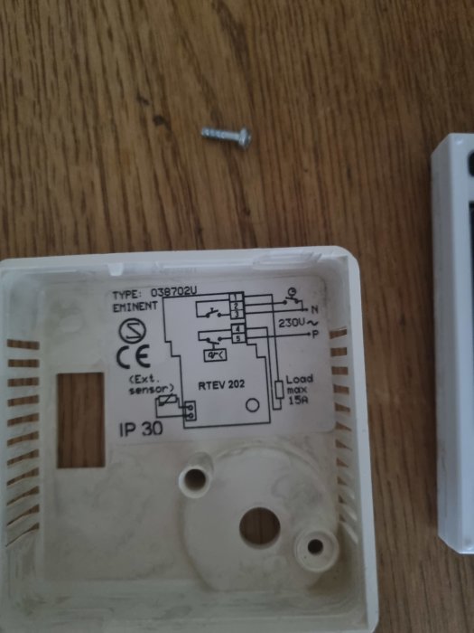 Bild på en borttagen golvvärmetermostat med elschema på baksidan och en skruv på trädgolvet.