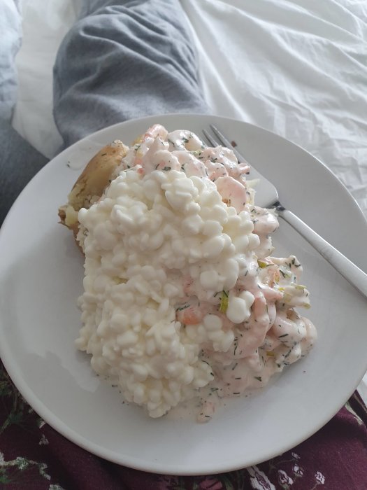 En tallrik med bakpotatis och rikligt med räkröra samt keso ovanpå, serverad på en säng.