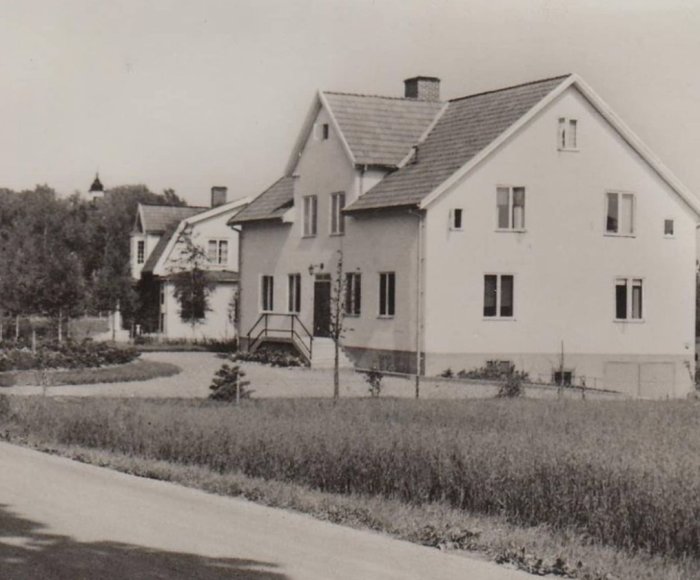 Svartvit bild av äldre hus med putsad fasad och parytterdörrar, omgivet av gräsmatta.